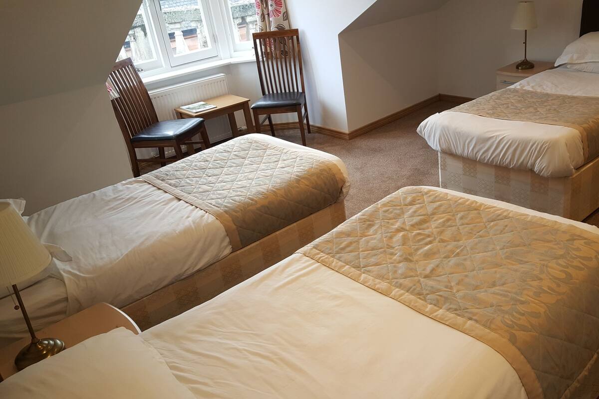 A hotel room with two single beds, a small table, a chair, and a window with curtains.