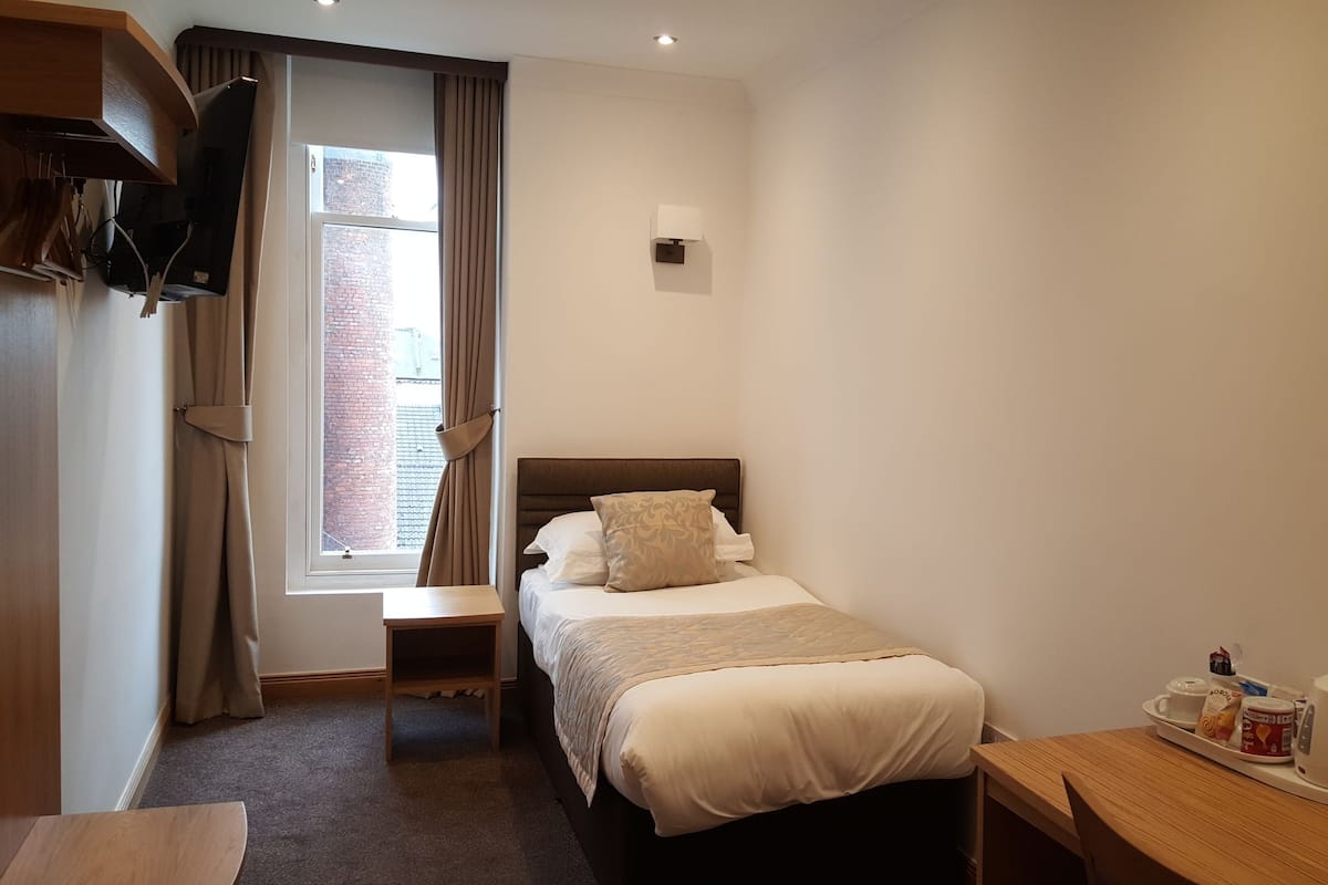 A hotel room with a bed, a bedside table with a kettle and tea set, a small table, a window with curtains, and a wall-mounted lamp.