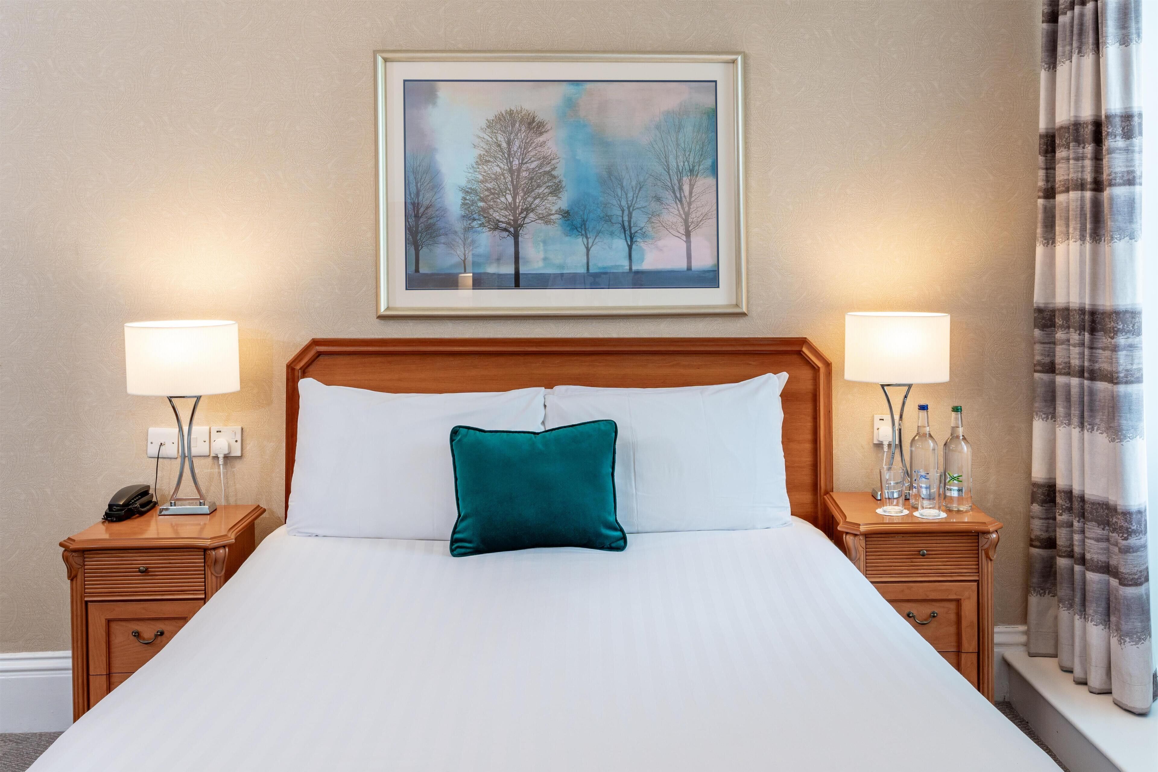 A hotel room with a large bed, two bedside tables, a nightstand, a painting above the bed, and a window with curtains.