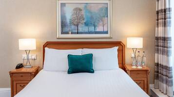 A hotel room with a large bed, two bedside tables, a nightstand, a painting above the bed, and a window with curtains.