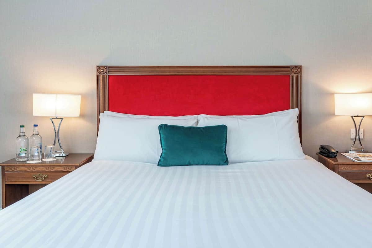 A hotel room with a large bed, two bedside tables, a red upholstered headboard, white bedding, and two bedside lamps.