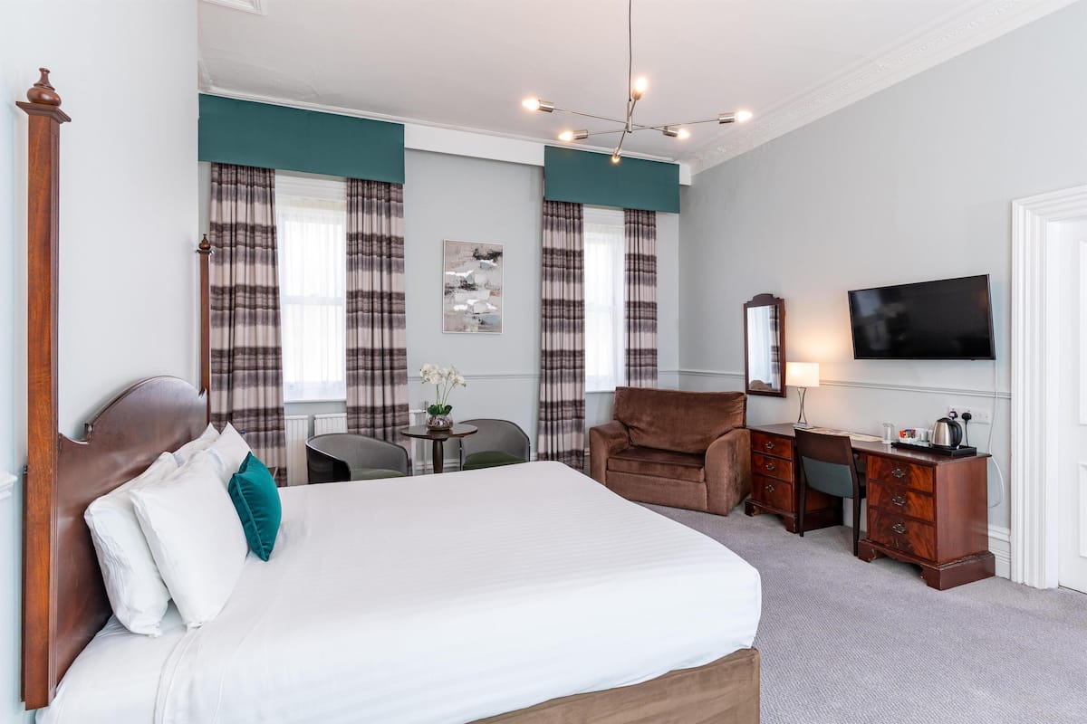 A hotel room with a large bed, a desk with a TV, a chair, and a small table with a lamp.