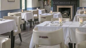 A dining area with tables set for four, each with white tablecloths and candles. There are chairs around the tables, and a fireplace is visible in the background.