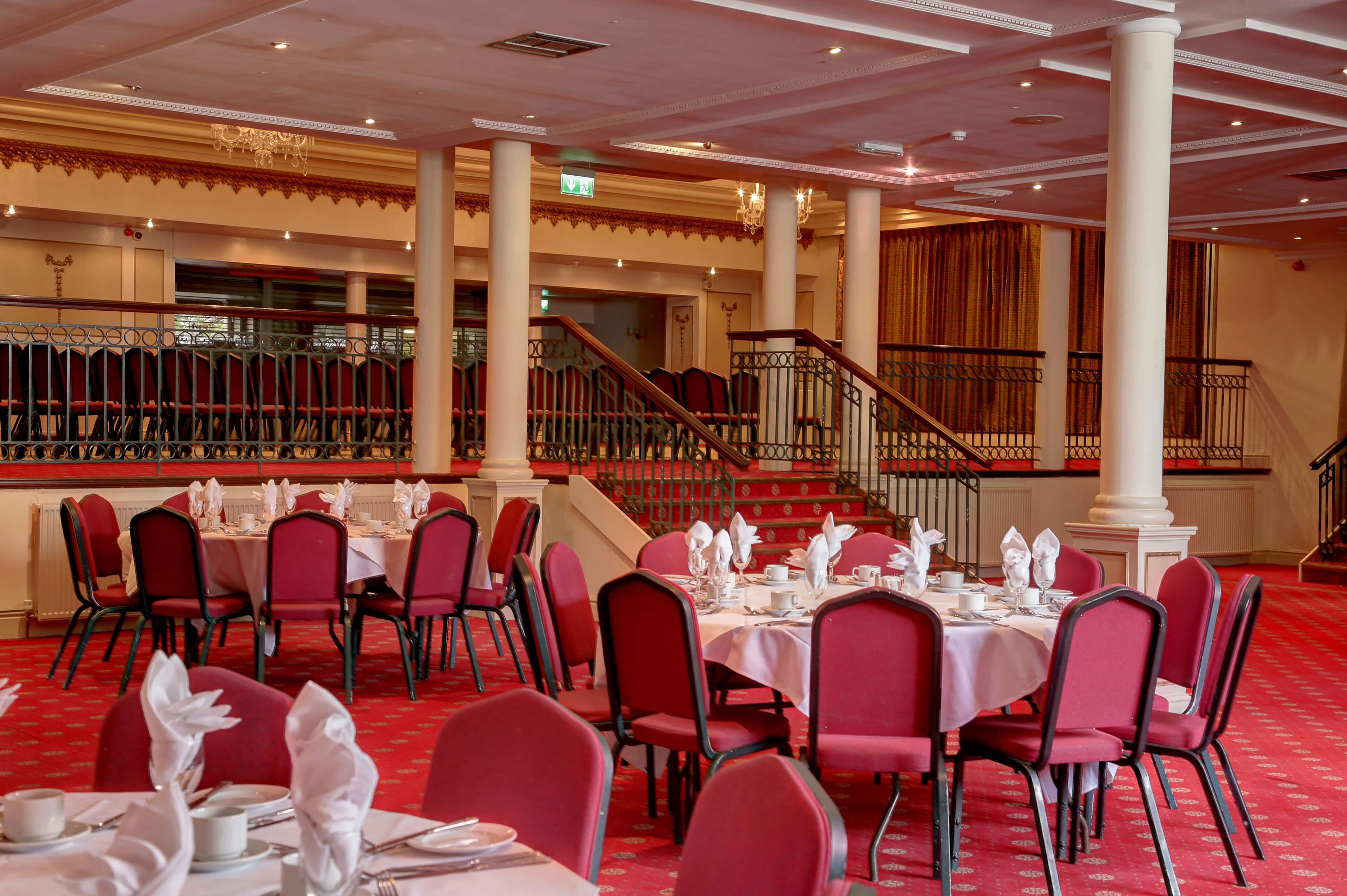 A banquet hall with tables set for dining, red chairs, and a staircase leading to an upper level.