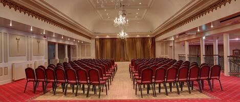 A conference room with rows of red chairs, a chandelier, and a red carpet.