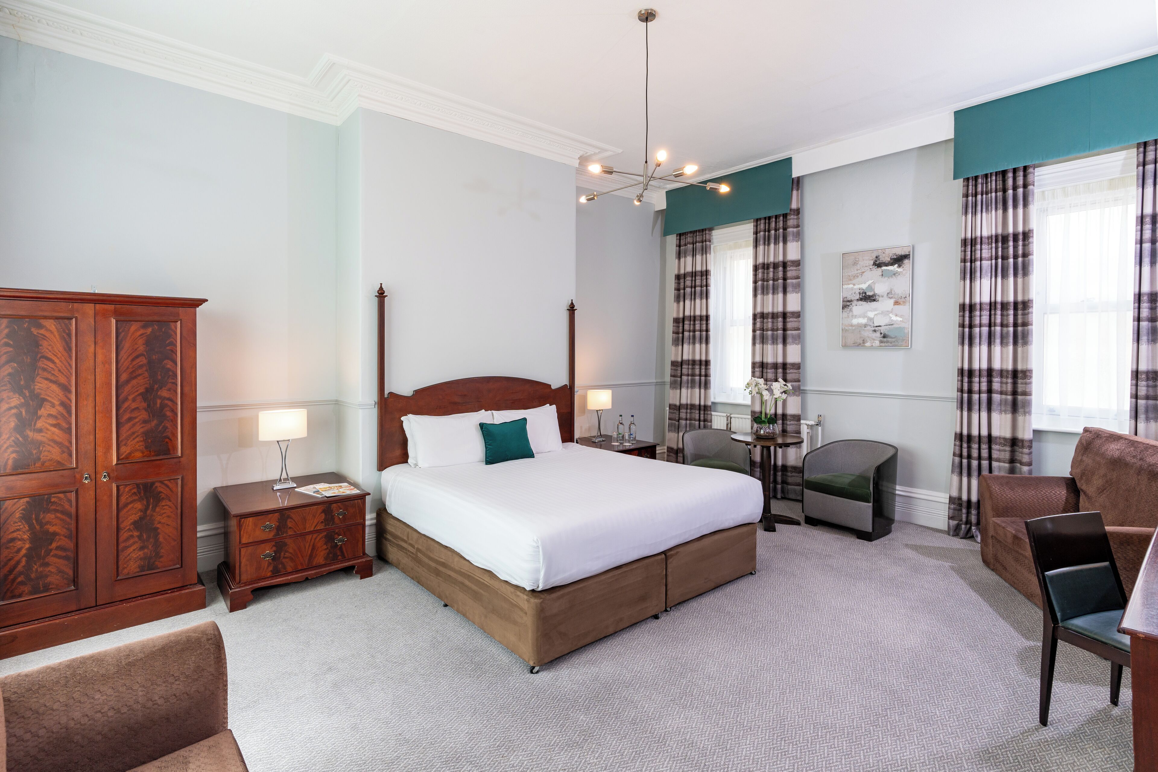 A hotel room with a large bed, a wooden wardrobe, a desk with a chair, and a sofa.