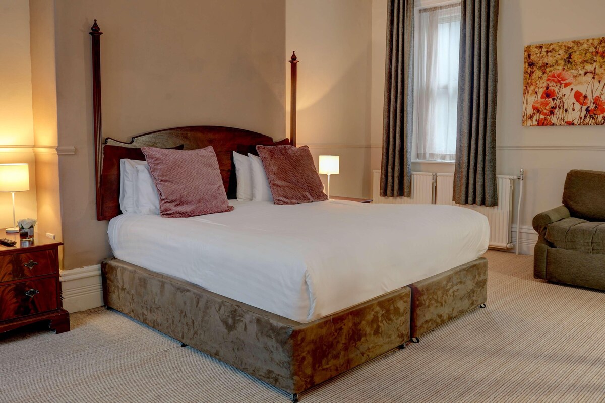 Ein Schlafzimmer mit einem großen Bett, einem Holzkopfende, einem Nachttisch mit Lampe, einem Sessel und einer Heizung.