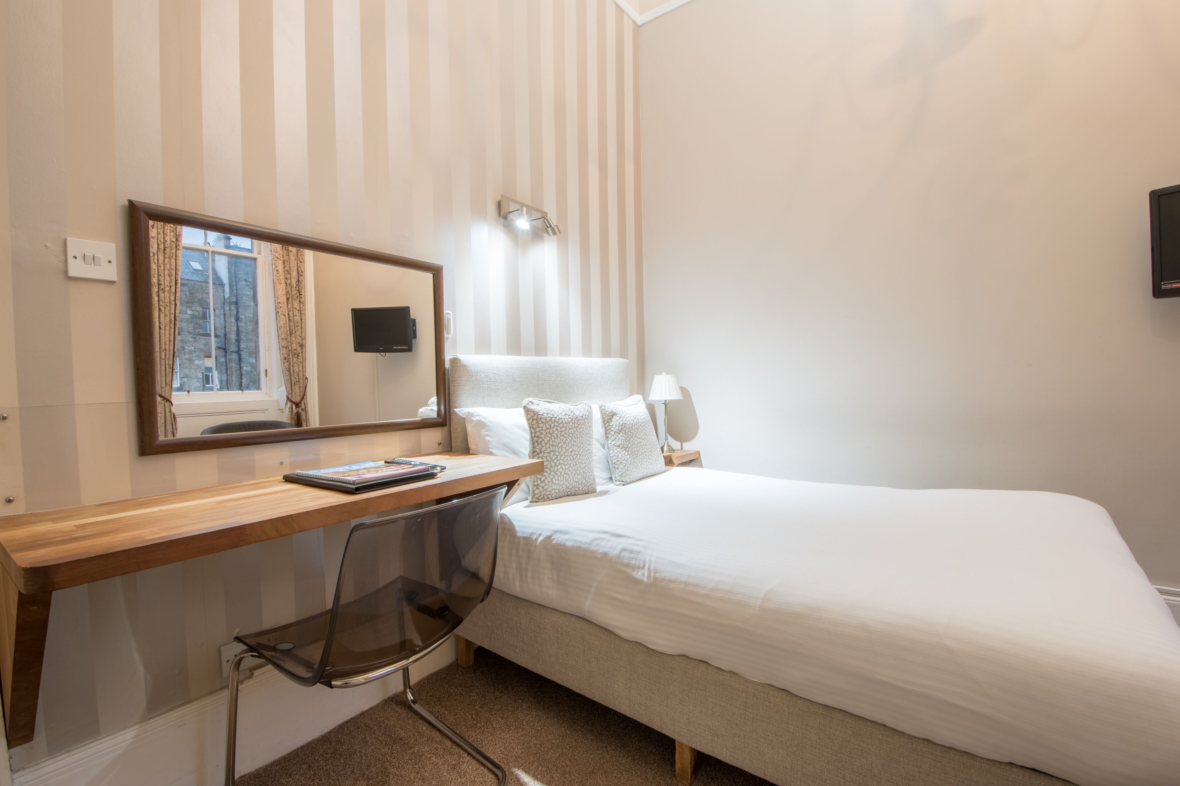 Hotellihuone, jossa on puinen työpöytä, tuoli, valkoisilla lakanoiden peitetty sänky, yöpöytä, lamppu, peili ja ikkuna, jossa on verhot.