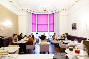Ein Restaurant mit einem zentralen, pinkfarbenen Fenster, weißen Wänden und einem Kronleuchter. Tische stehen um das Fenster herum, und Gäste speisen dort.