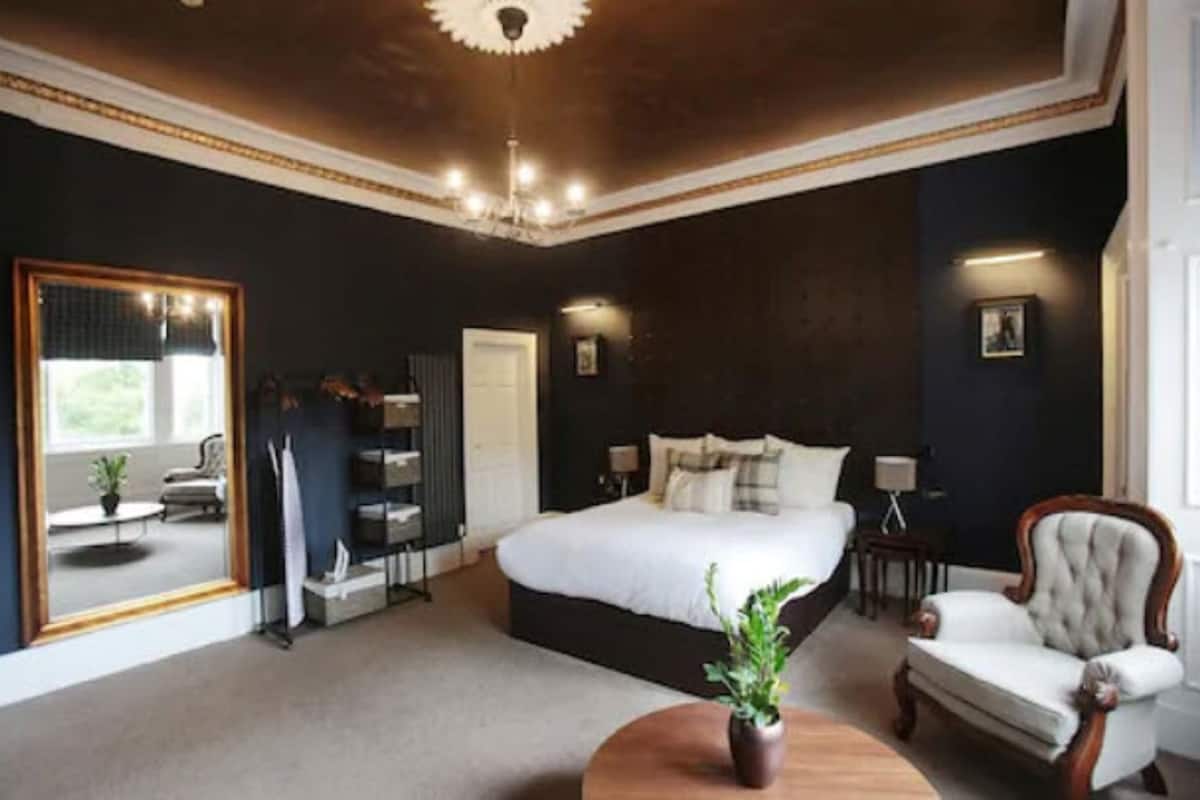 A bedroom with a large bed, a chandelier, a mirror, a chair, and a small table.