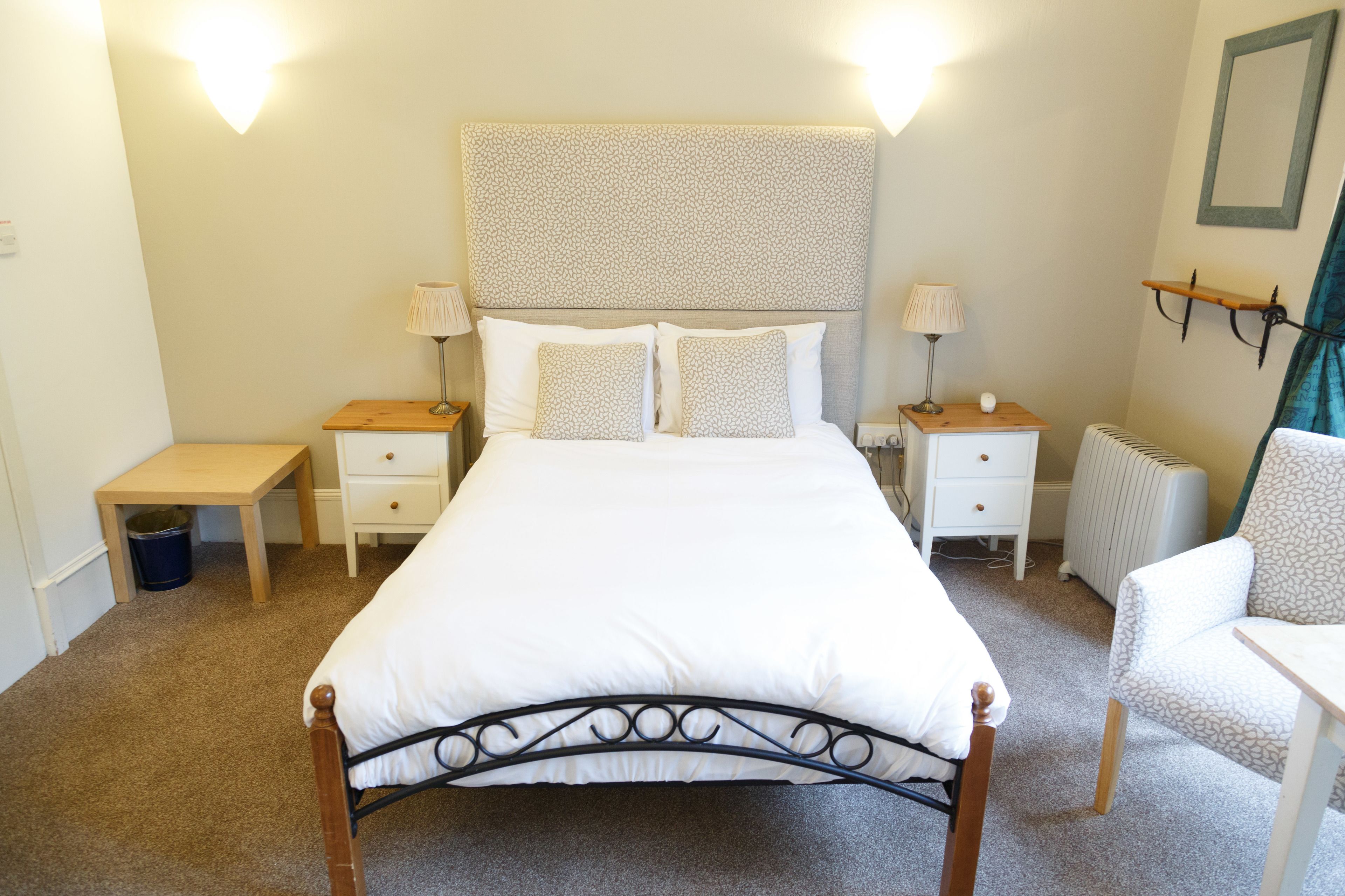 A bedroom with a bed, two bedside tables, a chair, a radiator, and wall-mounted lamps.