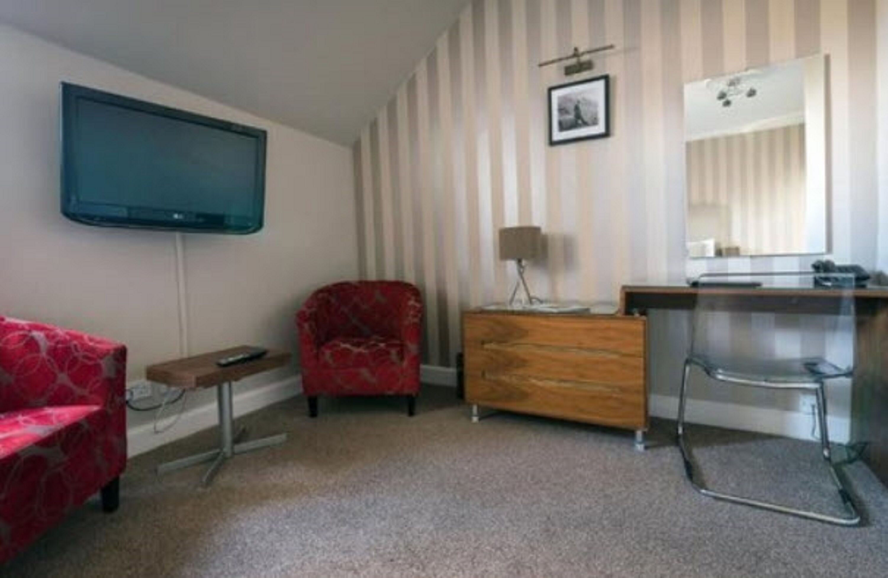 A room with a flat-screen TV mounted on the wall, a red armchair, a wooden dresser, a mirror, and a small table.