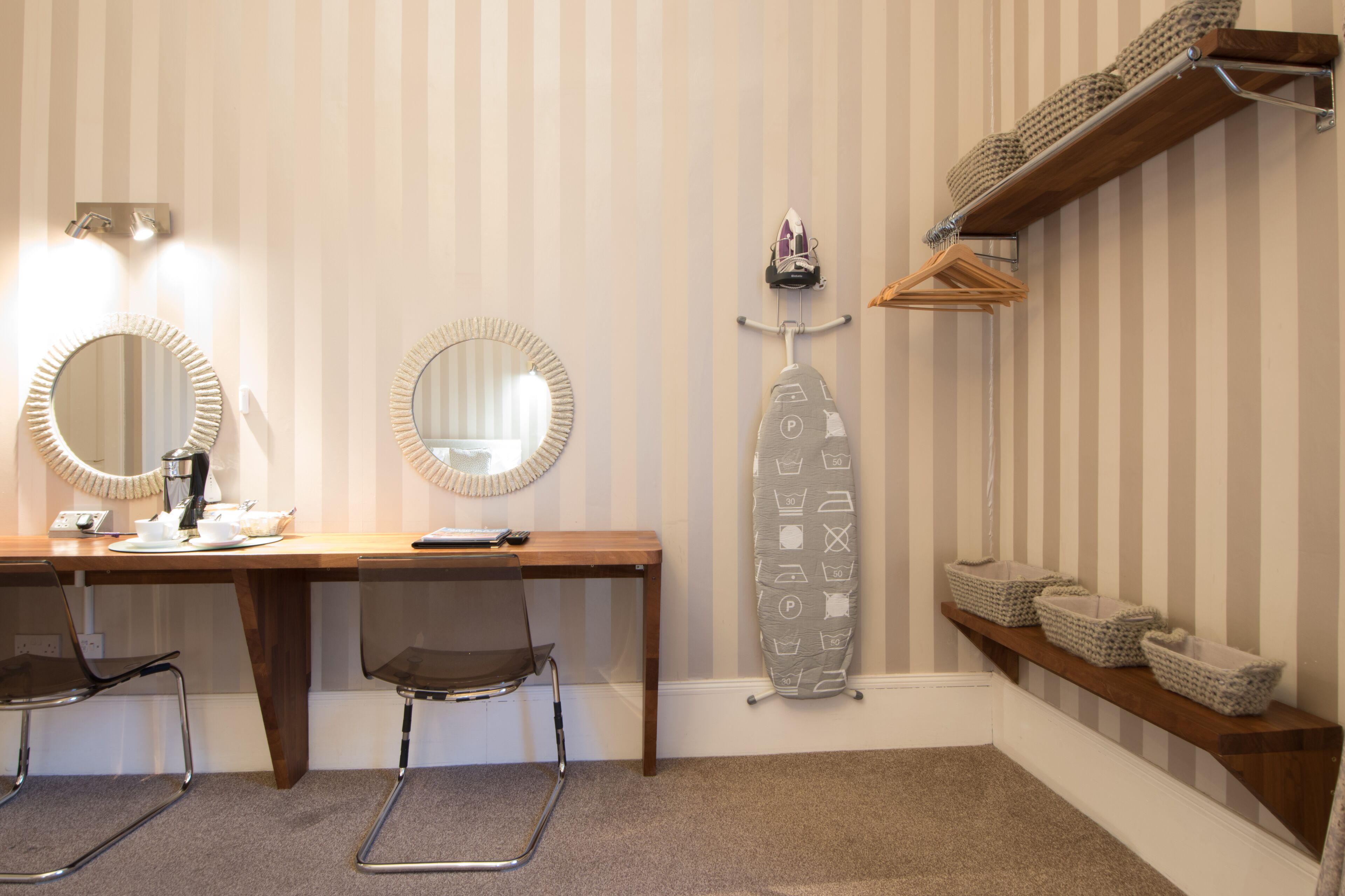 Una habitación con papel tapiz a rayas, un escritorio de madera con espejo, una silla y una estantería con cestas.