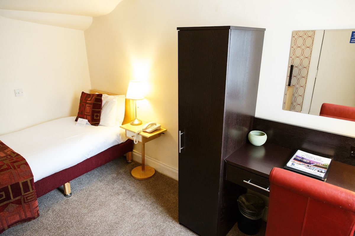 A hotel room with a bed, a desk with a lamp, a wardrobe, and a mirror.