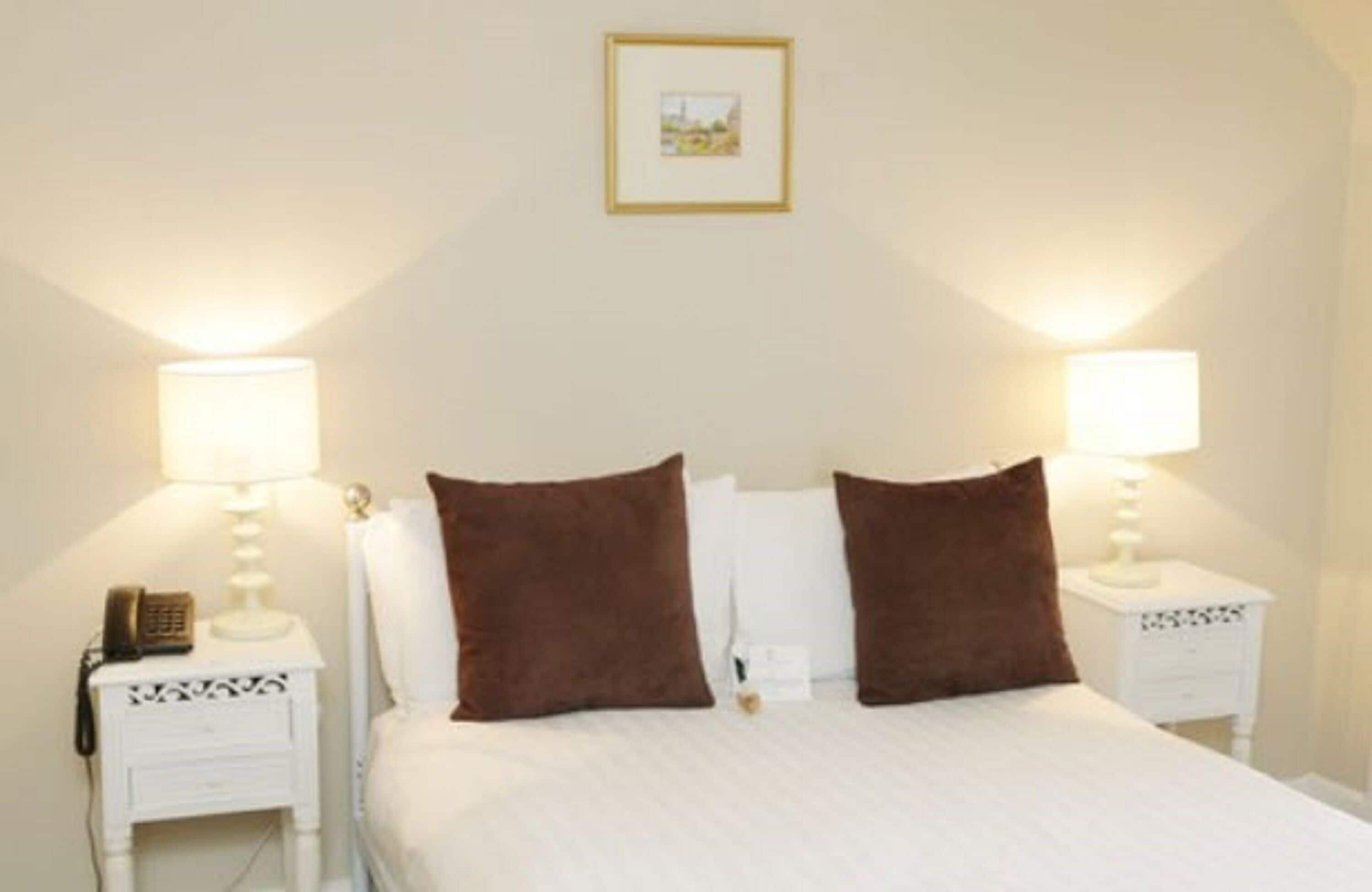 Een keurig opgemaakt bed met twee bruine kussens en witte beddengoed, met aan beide zijden nachtkastjes met lampen en elk een klein decoratief object.