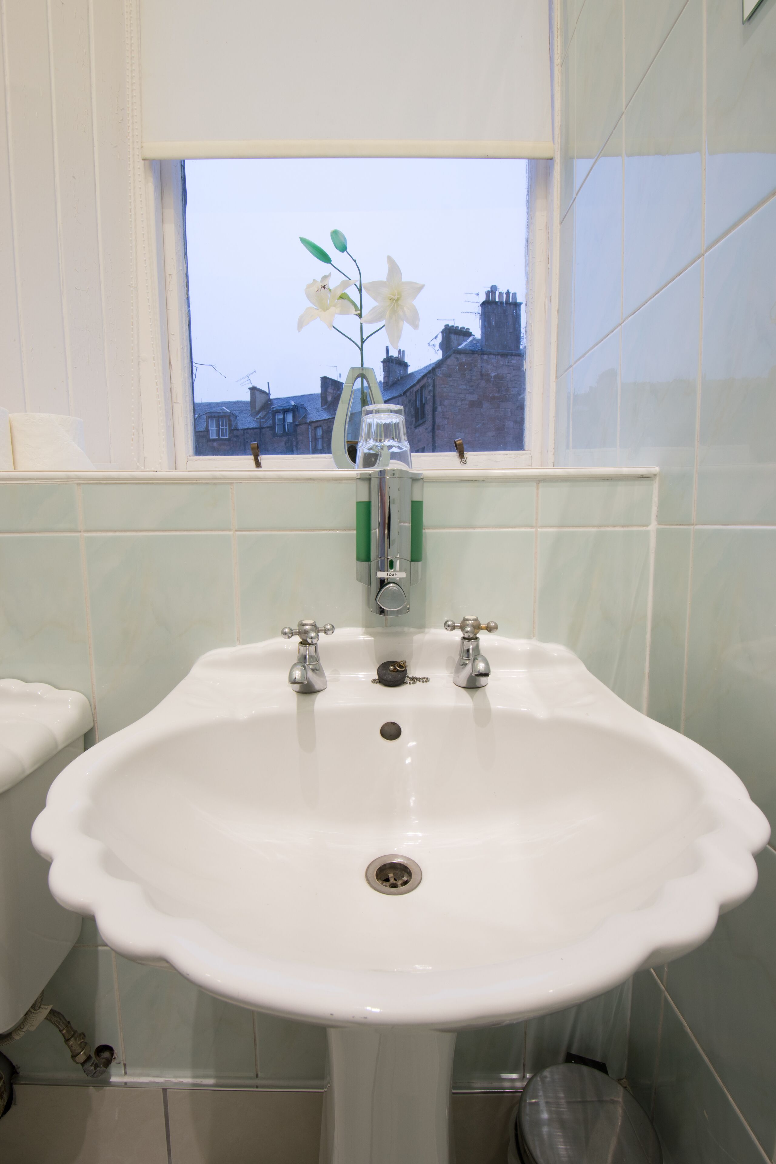 A bathroom with a white sink, two faucets, a window with a view of buildings, and a toilet.