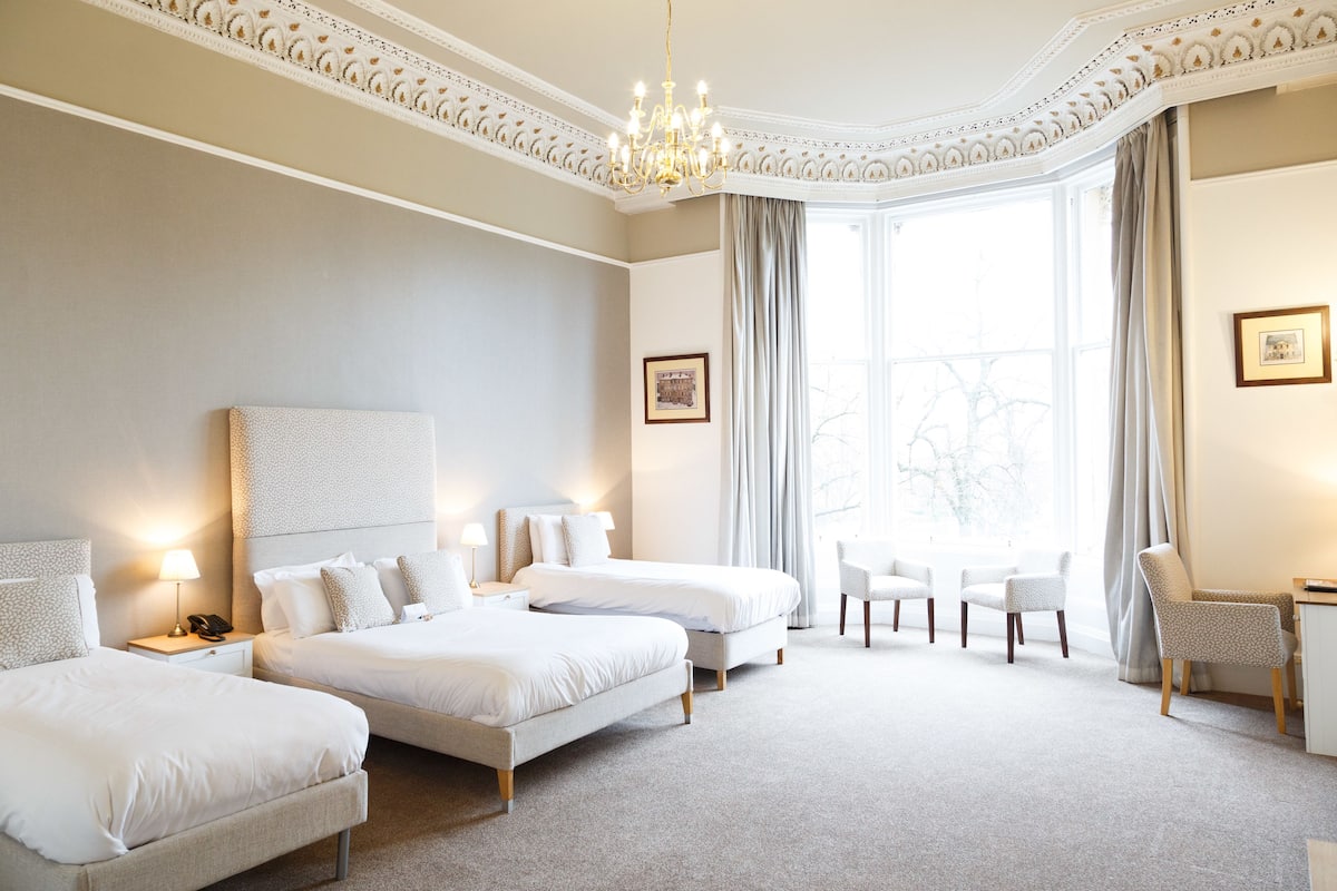 A hotel room with two beds, a chandelier, a large window with curtains, and a small table with a chair.