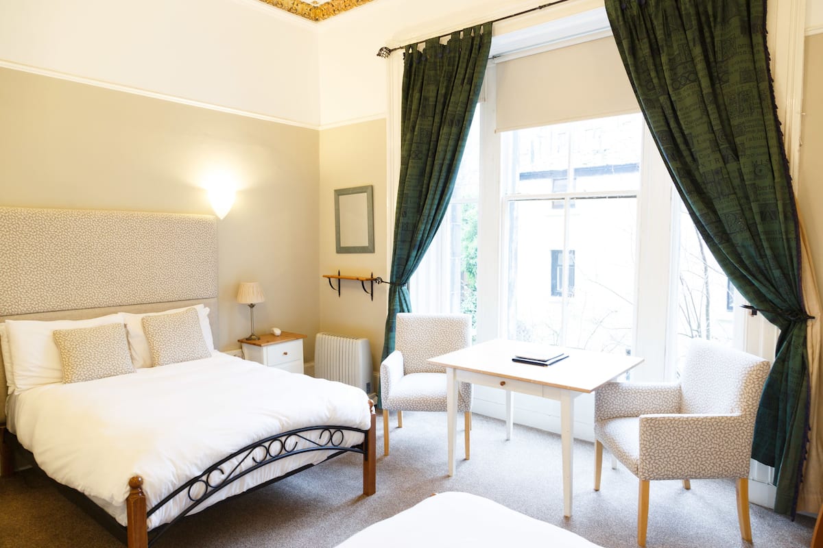 A bedroom with a bed, a desk, two chairs, a nightstand, and a window with curtains.