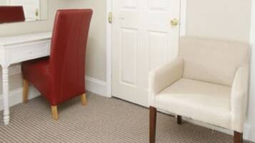A hotel room with a white desk, a red chair, a beige armchair, a television mounted on the wall, and a white door.