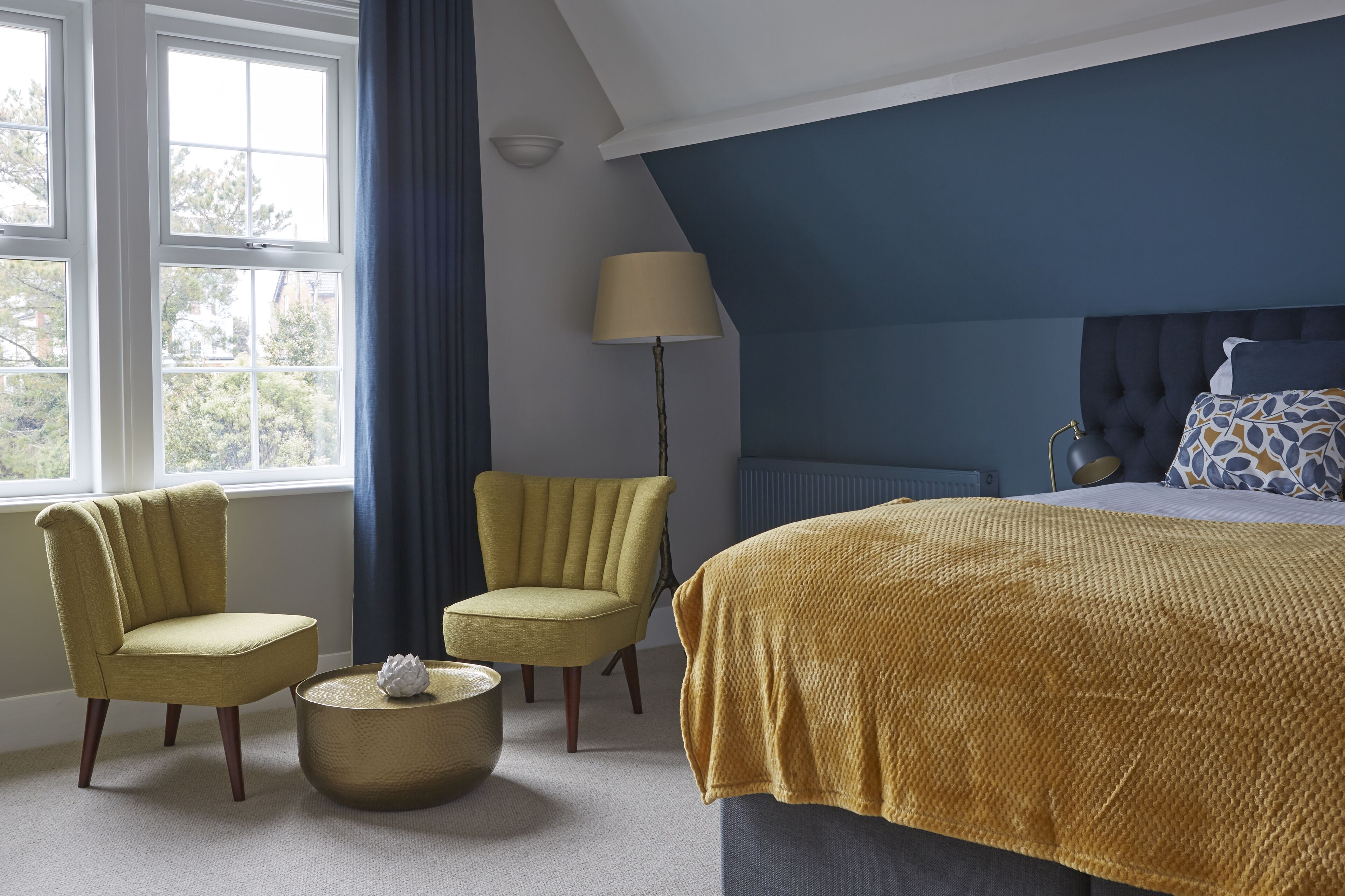 A bedroom with a bed, two armchairs, a round coffee table, a lamp, and a view of the outdoors through large windows.