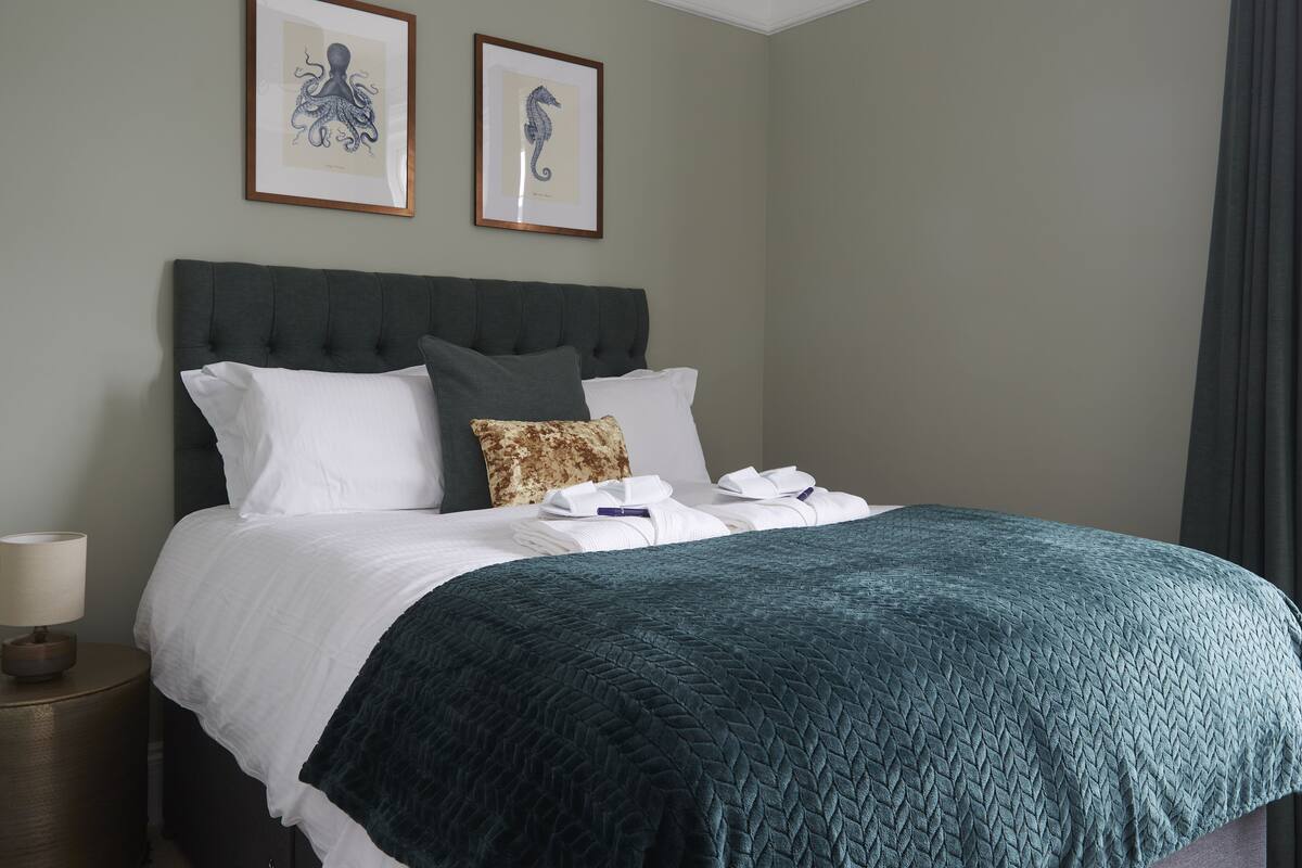 Cama bem arrumada com roupa de cama branca e verde escura, um travesseiro e uma toalha dobrada na cabeceira.