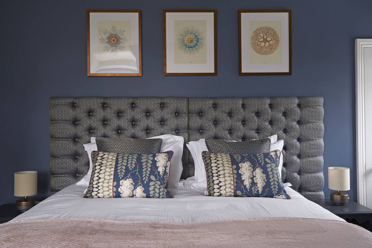 Cama bem arrumada com almofadas estampadas, cabeceira estofada e quadros na parede.