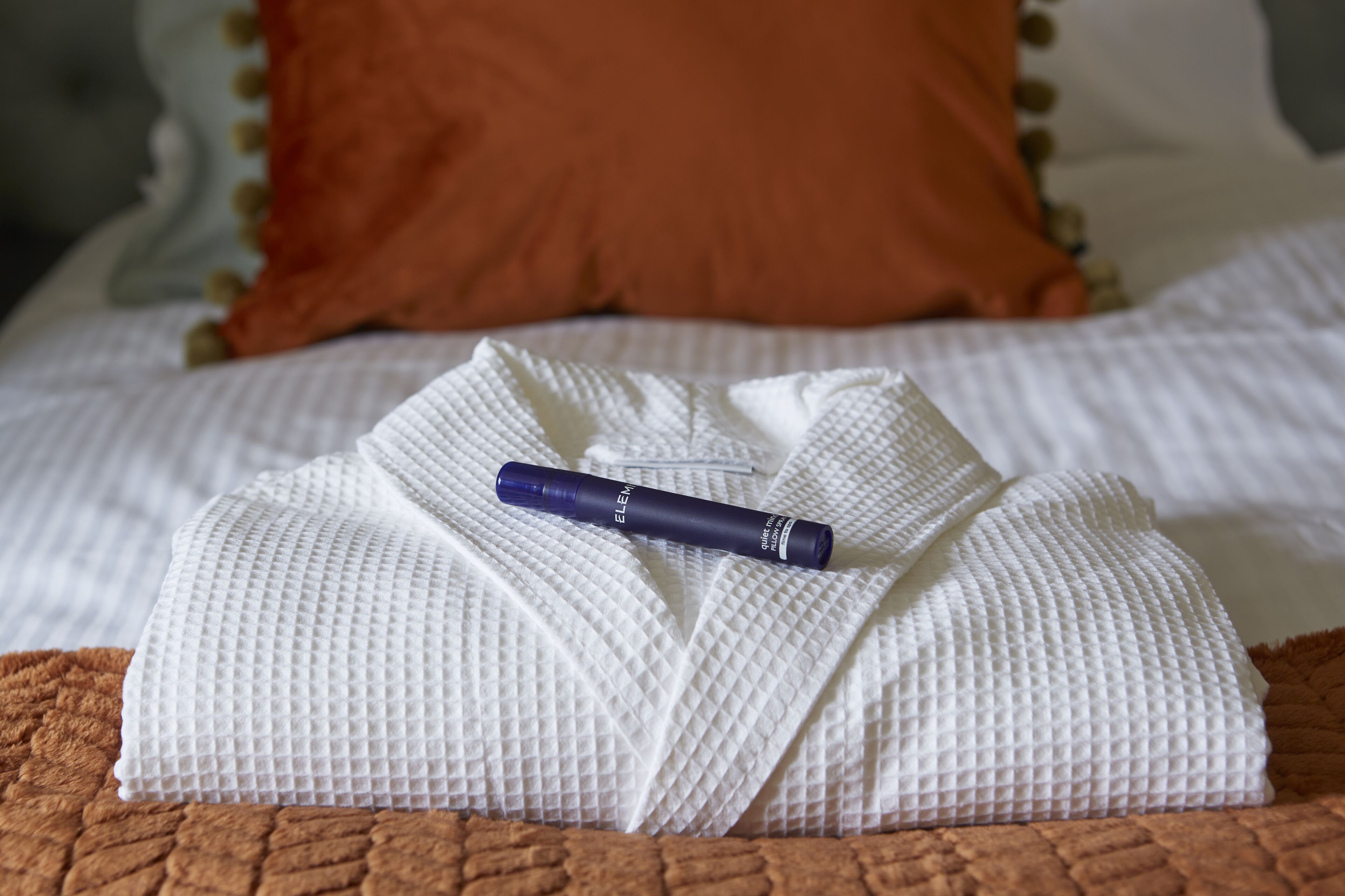 A neatly folded white bathrobe on a bed with a brown blanket and a blue bottle of Elem bath oil.