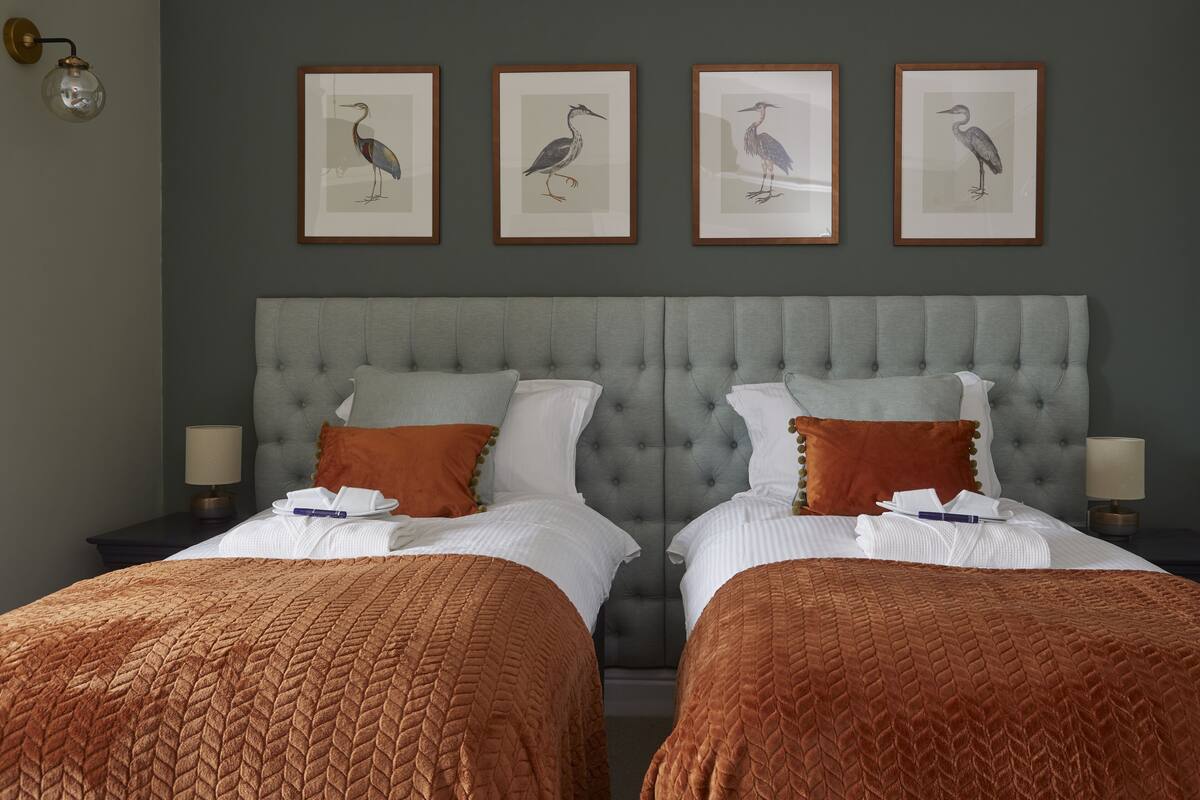Uma cama de casal com cabeceira estofada cinza, duas mesas de cabeceira com luminárias, e quatro ilustrações de pássaros emolduradas na parede.