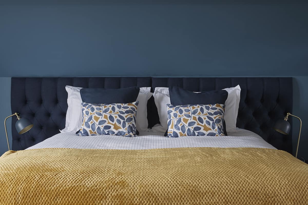 Cama bem arrumada com cabeceira escura, duas almofadas estampadas e um cobertor dourado.