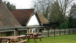 A building with a sloped roof, a wooden fence, and picnic tables in a grassy area. - The Roebuck Inn (Stevenage)