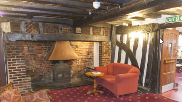 A cozy room with exposed brick walls, a fireplace, a red patterned carpet, and a single orange armchair. - The Roebuck Inn (Stevenage)