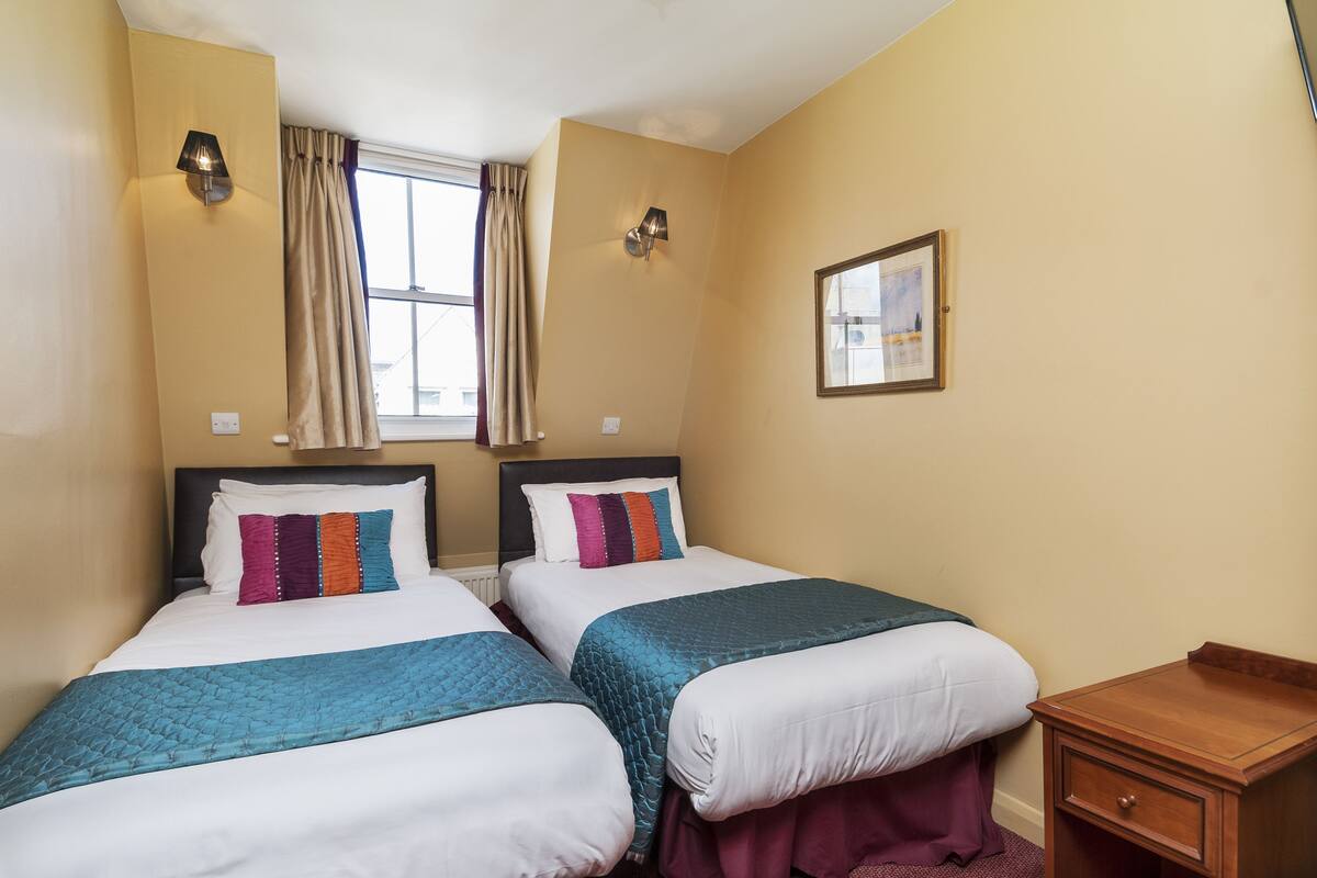 A hotel room with two beds, a window with curtains, a nightstand, and a framed picture on the wall.