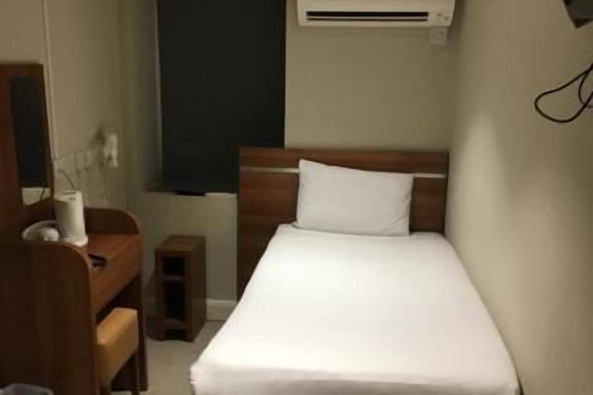 A single bed with a wooden headboard, a bedside table, a chair, a small stool, and a wall-mounted air conditioning unit.