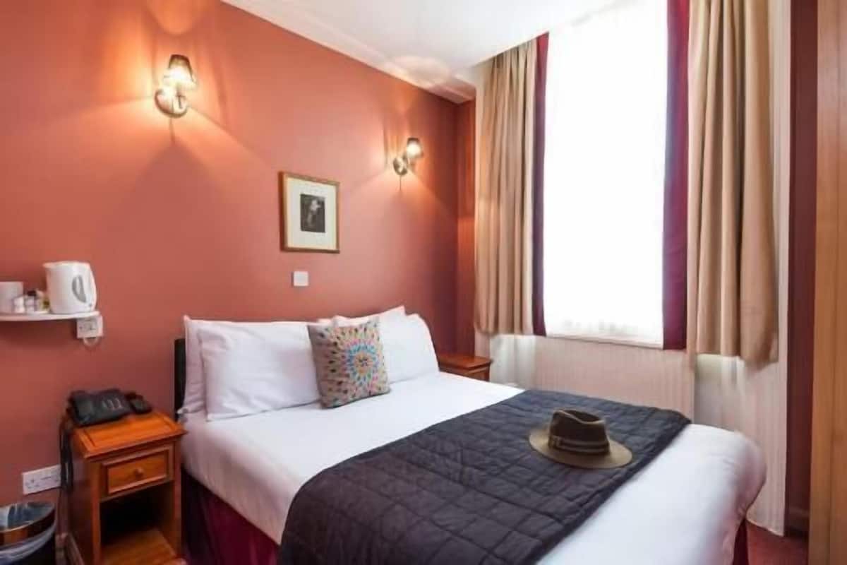A hotel room with a bed, bedside table, lamp, and a window with curtains.
