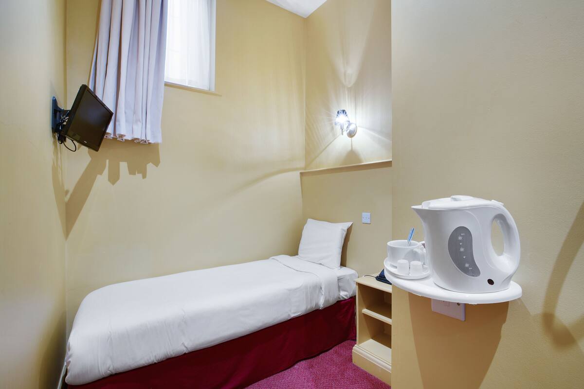 A small, single-bed room with a red bedspread, a white pillow, a wall-mounted TV, a kettle, and a cup on a shelf.