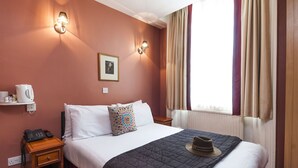 A hotel room with a bed, a bedside table with a phone, a nightstand, a wall-mounted lamp, a picture, and a window with curtains. - The Craven Hotel (London)