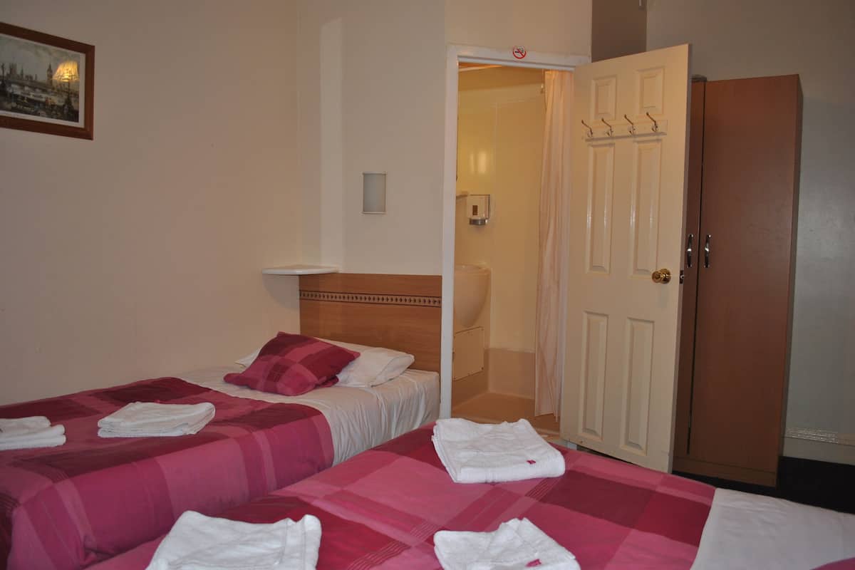 Une chambre d’hôtel avec deux lits, chacun recouvert d’une couvre-lit à carreaux roses et blancs, et des serviettes blanches pliées posées sur les lits. On y trouve une tête de lit en bois, une étagère blanche fixée au mur et une porte sur laquelle est accroché un manteau.