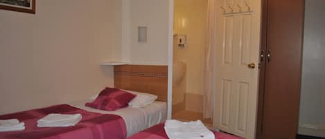 Habitación de hotel con dos camas, cada una con colcha a cuadros rosa y blanca, y toallas blancas dobladas sobre ellas. Se observa un cabecero de madera, un estante blanco montado en la pared, y una puerta con un abrigo colgado en el pomo.