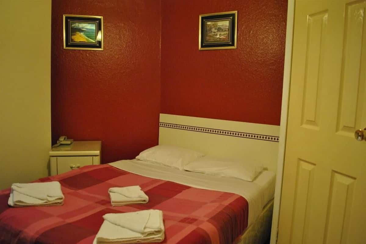 Une chambre d’hôtel avec un mur rouge, un lit avec une literie blanche, deux tableaux encadrés au mur et une porte blanche.
