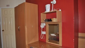 Ein Zimmer mit Holzschrank, Schreibtisch mit Stuhl, Spiegel, Fön und einem Regal mit diversen Gegenständen.