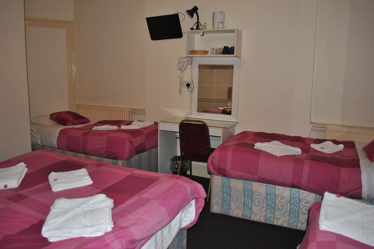 Une chambre d’hôtel avec trois lits, chacun recouvert d’une couverture rouge et blanche, et des serviettes blanches pliées posées sur les lits. On y trouve un bureau avec un écran d’ordinateur, une lampe et un téléphone.