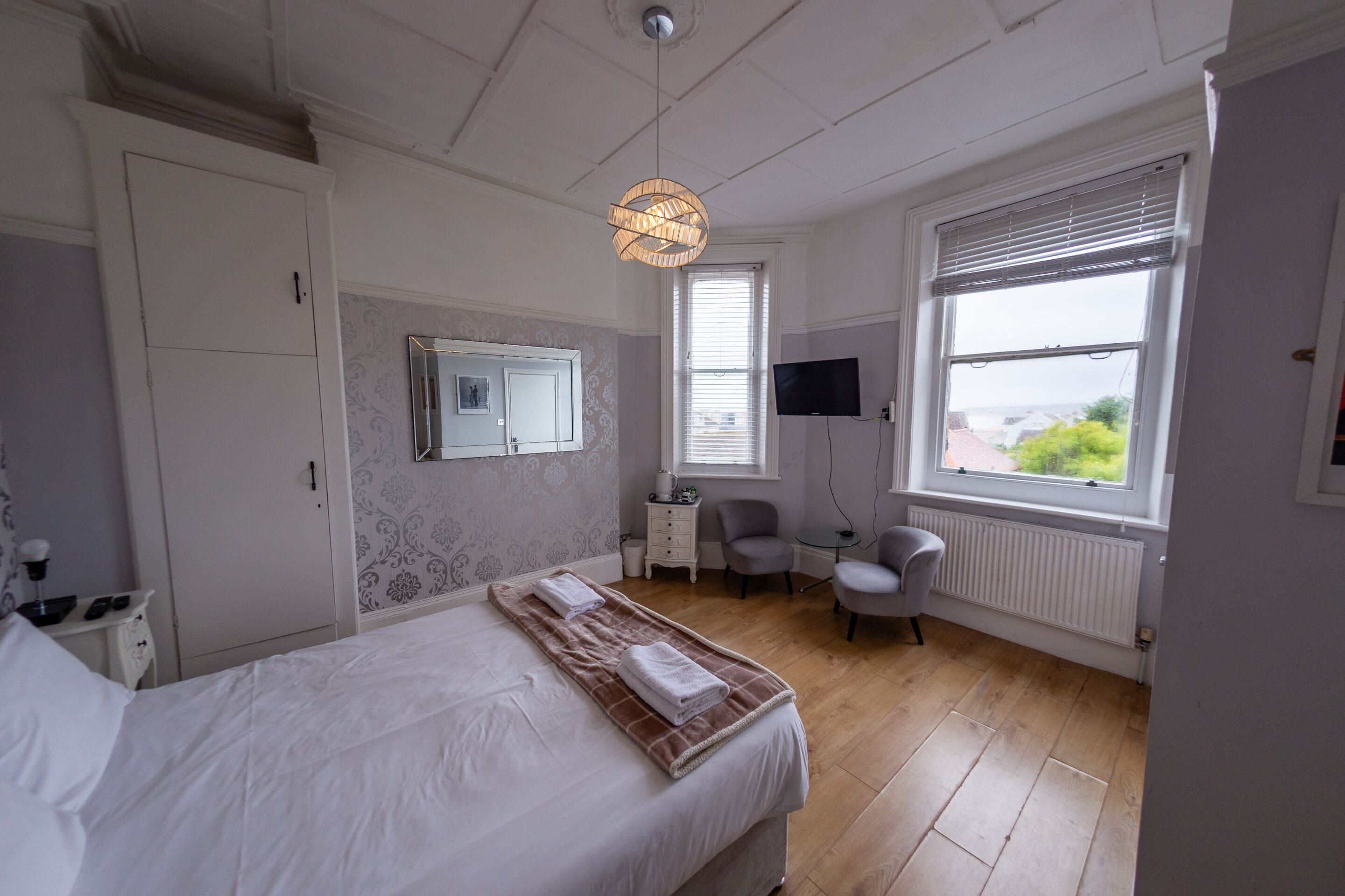 Un dormitorio con cama, mesita de noche, una silla, televisor y ventana con persianas.