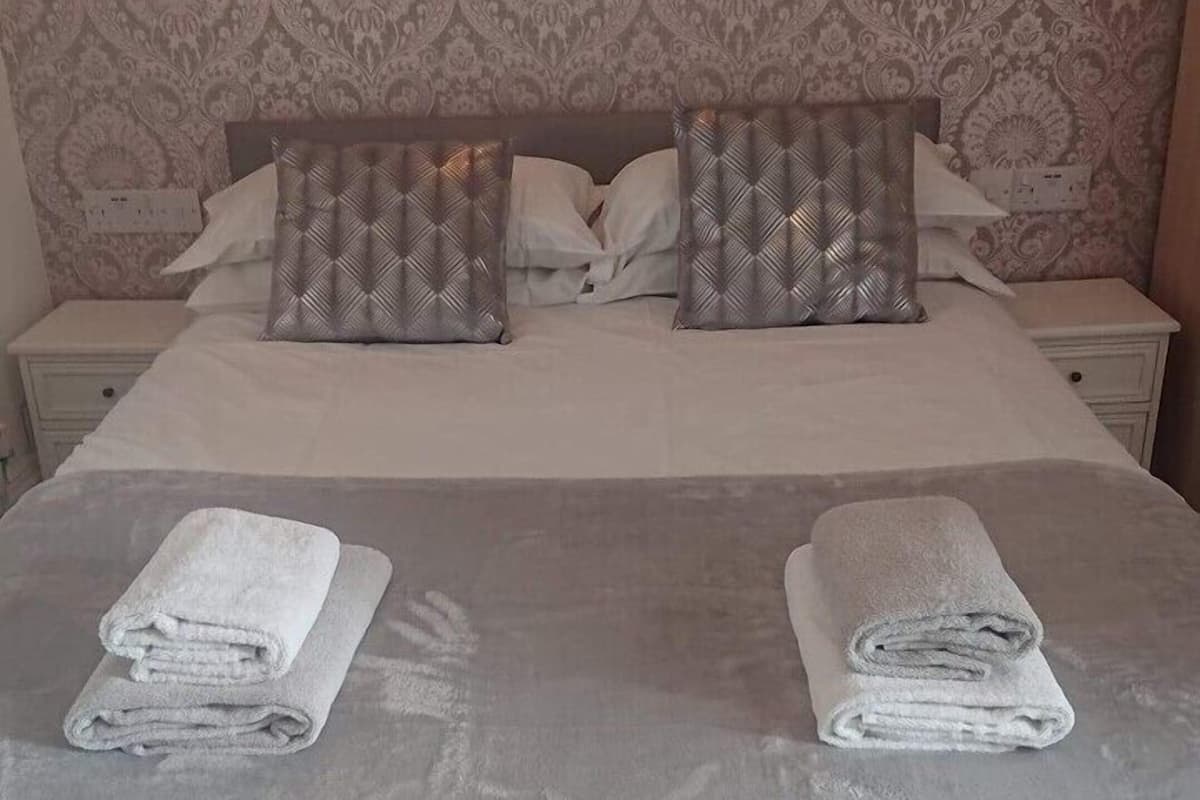 Un lit bien fait, avec deux oreillers, une table de chevet et un mur recouvert d’un papier peint à motifs.