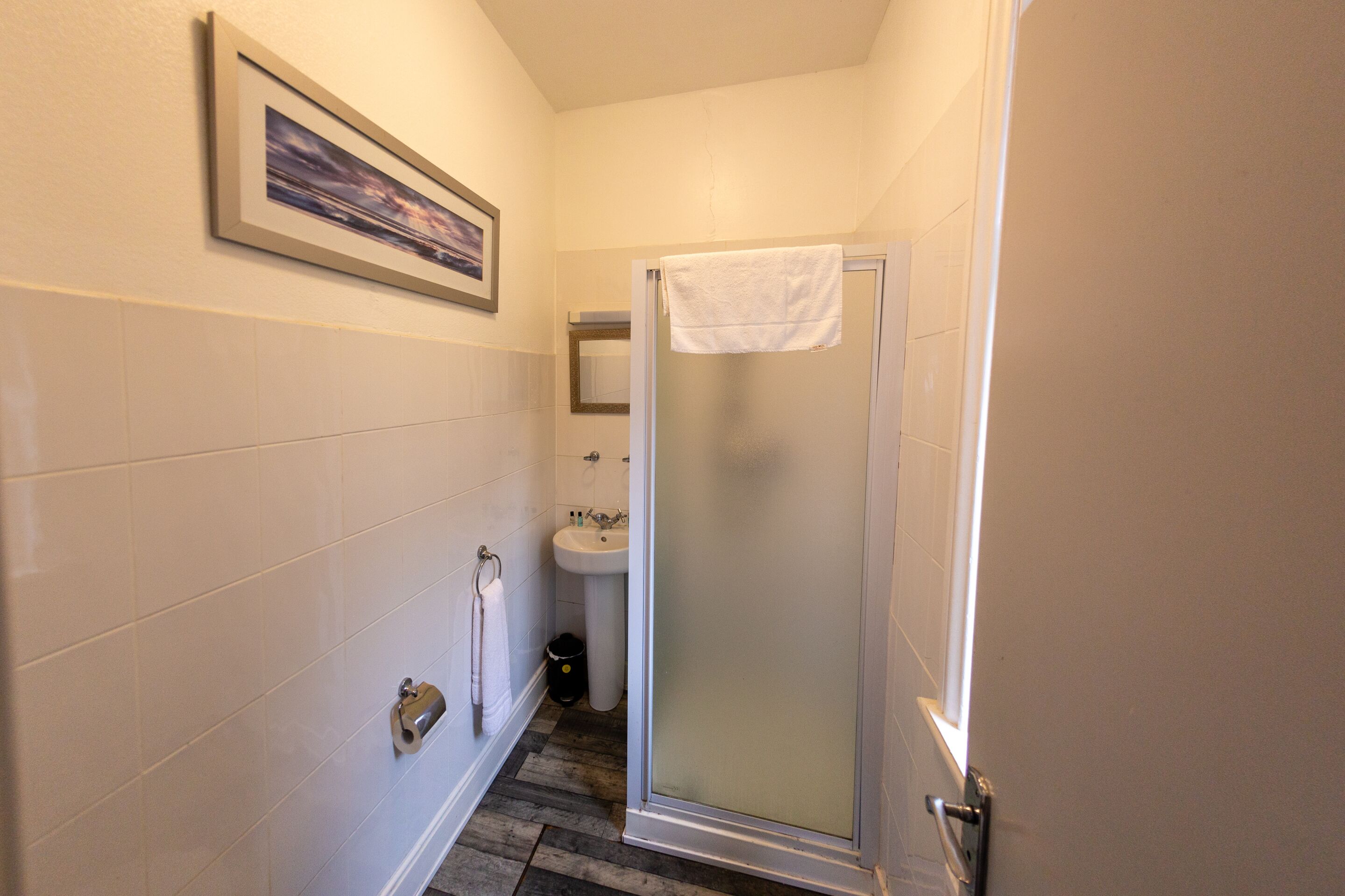 A bathroom with white tiled walls, a framed picture, a sink, and a glass-enclosed shower.