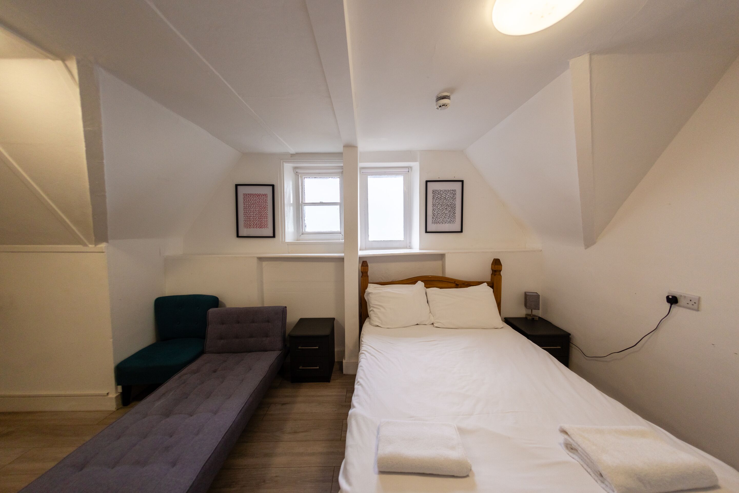 Ein Schlafzimmer mit einem Bett, einer Bank, einem Nachttisch, einer Lampe und einem Fenster.