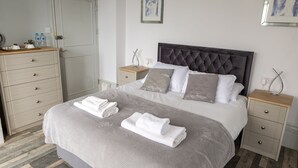 Un lit bien fait, recouvert d’une couverture grise et agrémenté de deux oreillers, une table de chevet avec une lampe, et une commode en bois sur laquelle est accrochée une photo encadrée au mur.