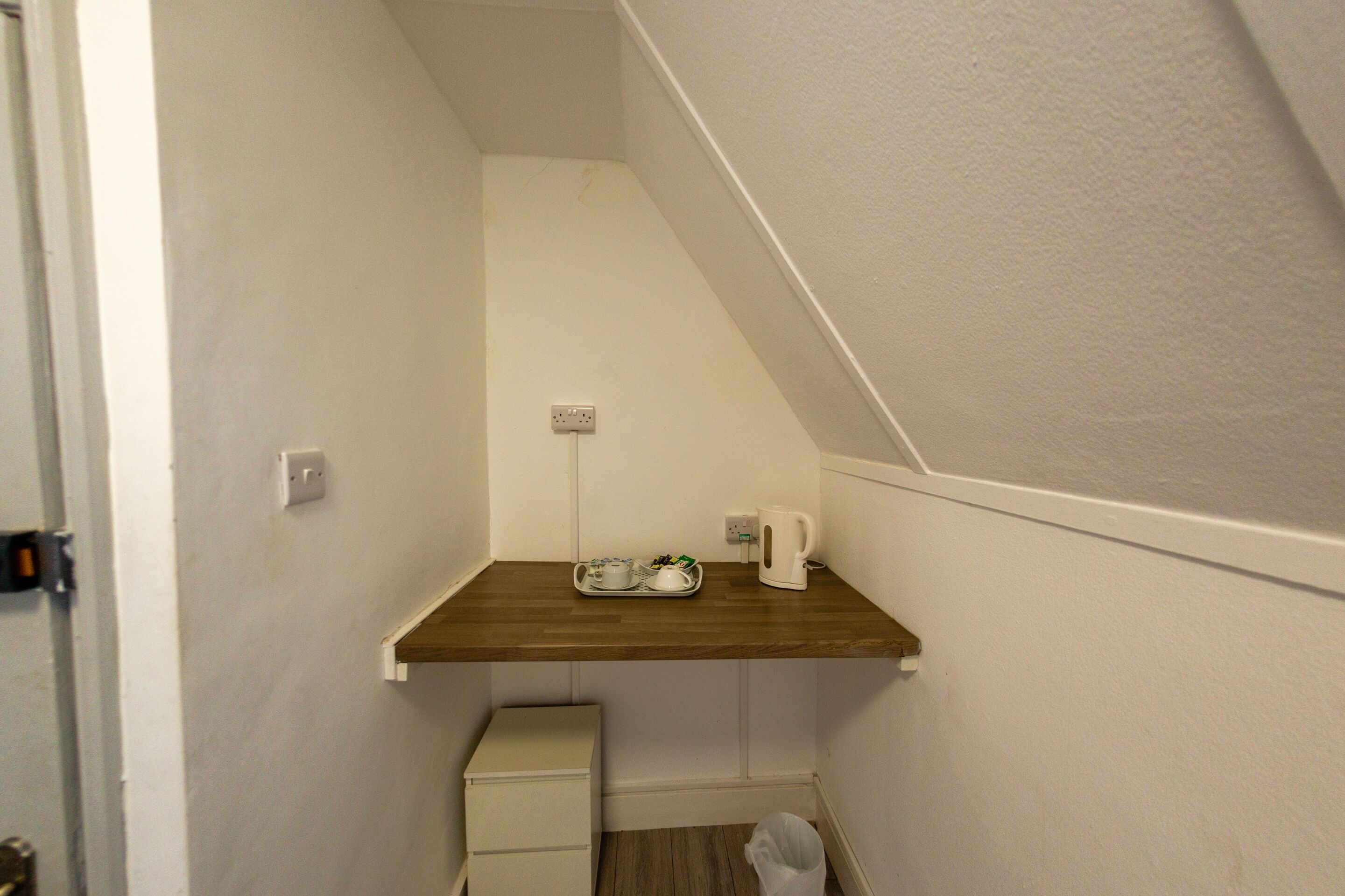 A small, narrow room with a wooden shelf, a white cabinet, and a trash bin.