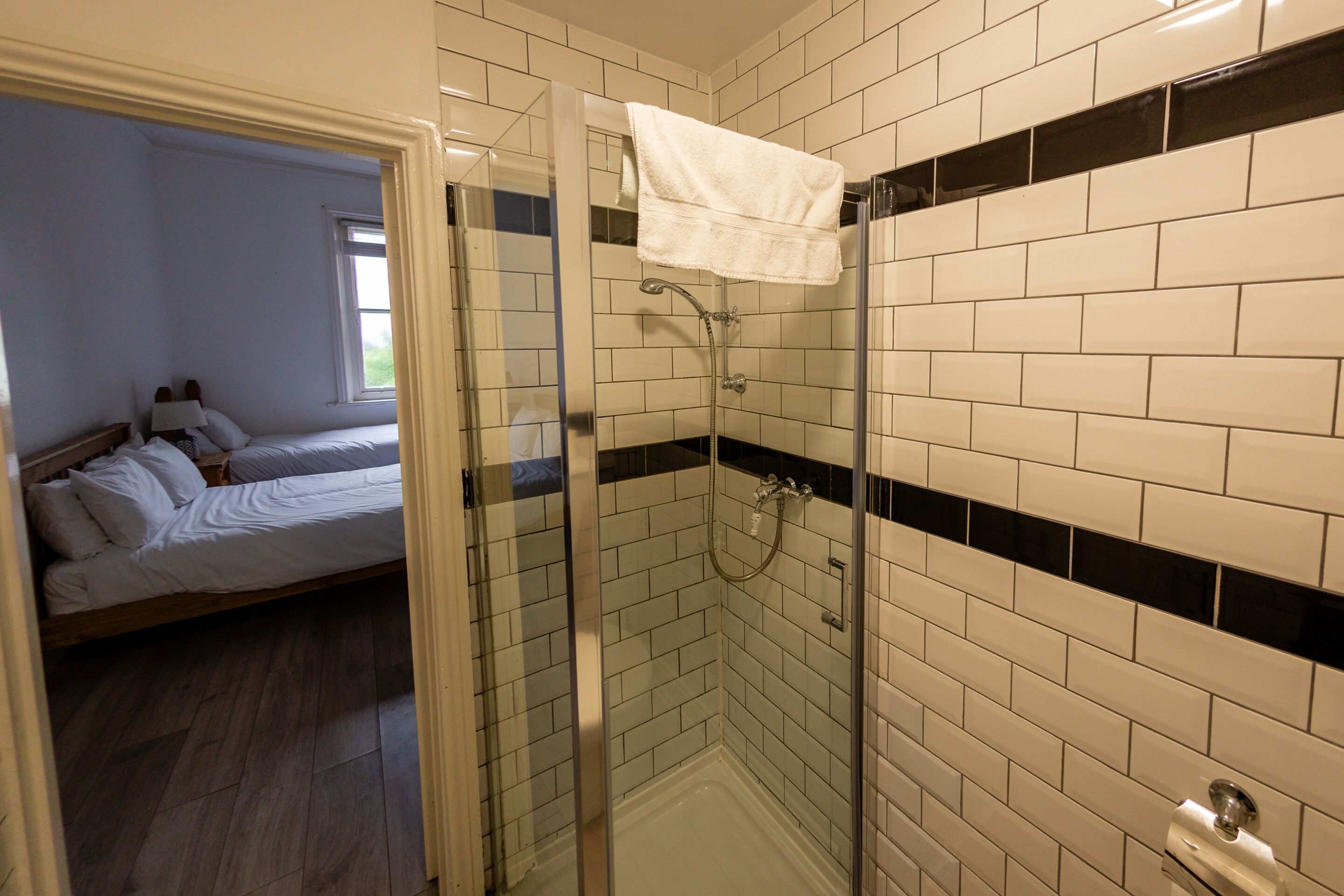 Un bagno con doccia vetrata, pareti piastrellate di bianco e un letto nella stanza adiacente.