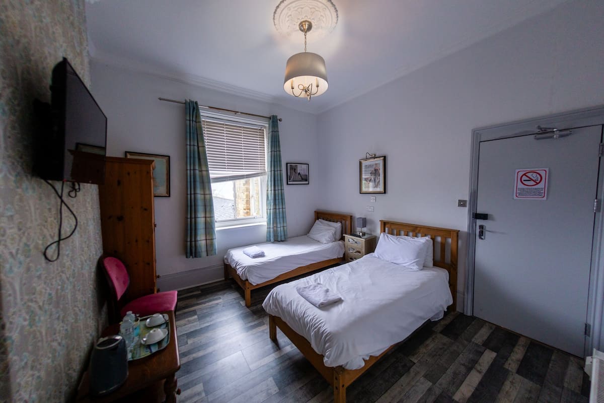 Une chambre d’hôtel avec deux lits simples, une armoire en bois, une télévision, une fenêtre avec des rideaux et une porte sur laquelle est apposé un panneau « interdiction de fumer ».