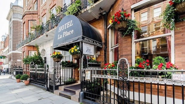 Fachada de un hotel con un toldo negro, jardineras con flores y un letrero que dice "HOTEL La Place".
